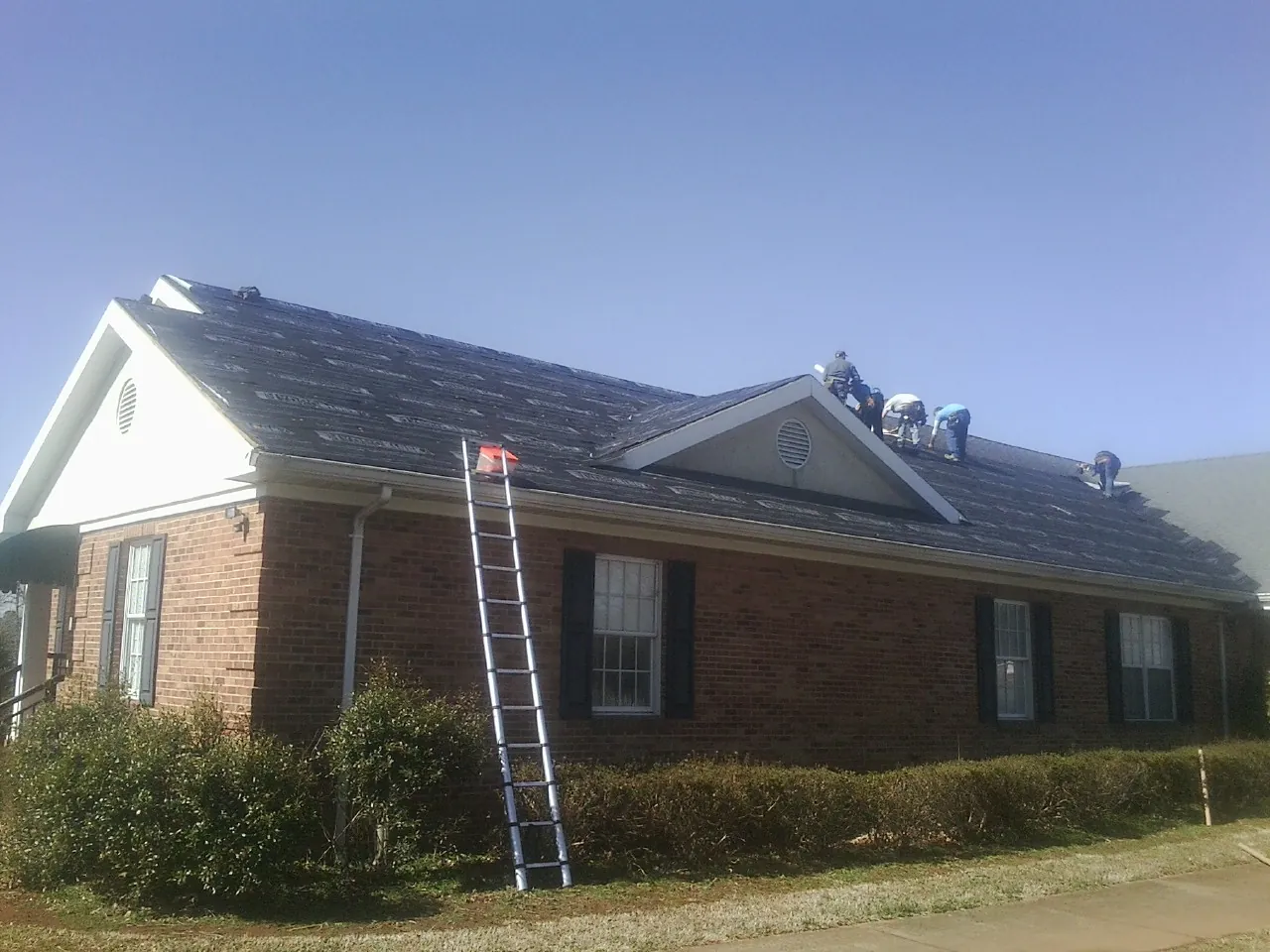 Crew tearing shingles off a brick ranch roof with a ladder set — Emergency Roof Repair work by our crew in Huron